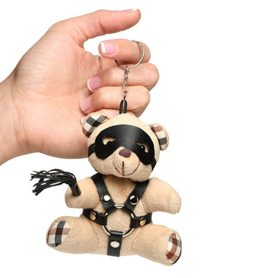 Hand holding a small beige teddy bear keychain wearing a black faux leather harness, eye mask, and holding a mini flogger.