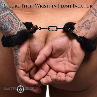 Close-up of tattooed wrists secured in black furry handcuffs with a connecting chain against bare skin background.