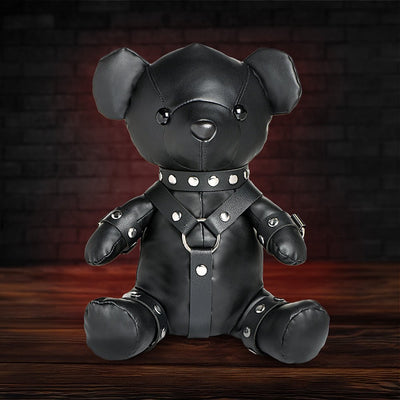 Black vegan leather teddy bear dressed in detailed gimp harness with silver studs, sitting on wooden surface against dark background.
