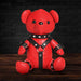 Red vegan leather teddy bear wearing black studded gimp harness and cuffs, sitting on wooden surface with dark background.