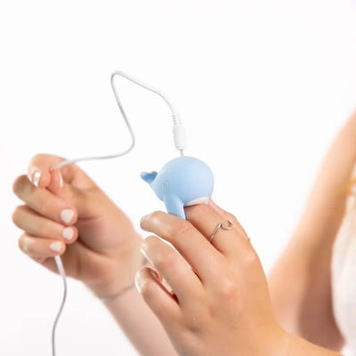 Hand holding and charging a blue whale-shaped finger vibrator with a white USB cable.