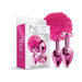 Nixie metal butt plug set in pink metallic with a fluffy pom pom and a jewel-inlaid base, displayed with packaging box.