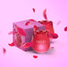 Red rose-shaped Nuud pulsing vibrator with packaging and floating rose petals on a pink background.
