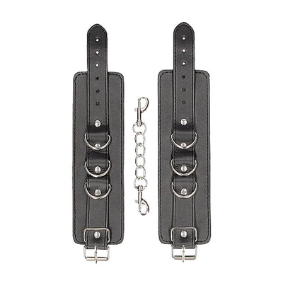 Black bonded leather wrist or ankle cuffs with adjustable straps and metal chain connector on white background.
