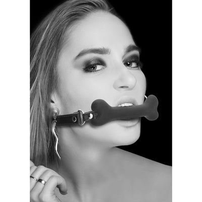 Black and white photo of a woman wearing a black silicone bone-shaped gag with adjustable bonded leather straps.