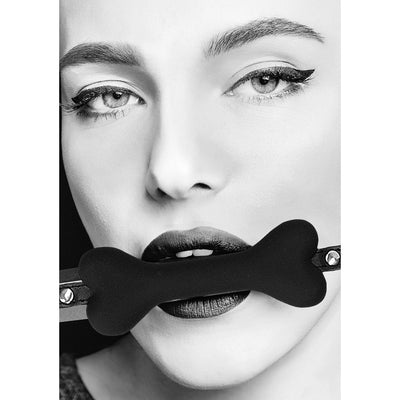 Close-up black and white photo of a person wearing a black silicone bone-shaped gag with adjustable leather straps.