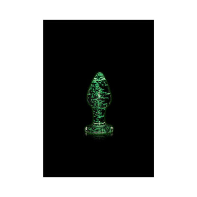 Clear glass butt plug glowing green in the dark with speckled pattern and smooth tapered shape on black background.
