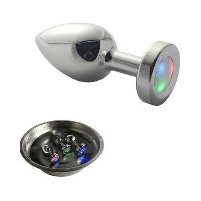 Small polished metal anal plug with LED light-up base showing red, green, and blue colors, displayed with three plugs in water bowl.