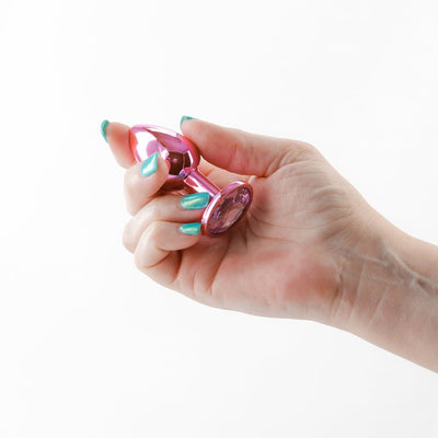 Hand holding a small, shiny pink metal anal plug with a jeweled base against a white background.