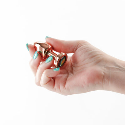 Hand holding a petite rose gold metal anal plug with a rainbow-colored gem base against a white background.