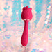 Red rose-shaped clitoral and G-spot vibrator displayed on a clear stand with a pastel bokeh background.