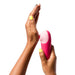 Hand holding Romp Shine X Pink silicone clitoral stimulator with yellow nail polish on white background