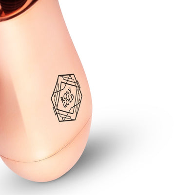 Close-up of the rosy gold mini massager with geometric logo on a smooth metallic surface against a white background.