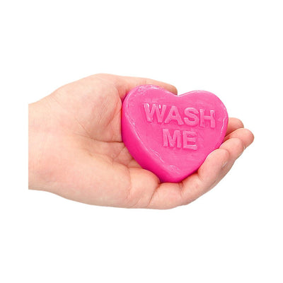 Hand holding a bright pink heart-shaped soap bar embossed with the words "WASH ME" on a white background.