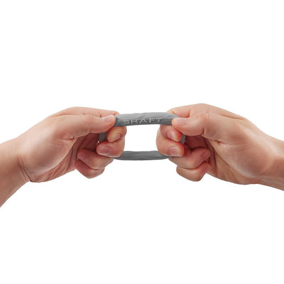 Hands stretching a small gray flexible silicone Shaft C-ring with embossed logo on white background.