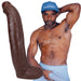 Man wearing a blue cap and light blue shorts posing next to a large lifelike chocolate-colored dildo with textured veins and suction cup base.