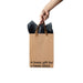Hand holding a kraft paper gift bag with brown ribbon handles and black tissue paper, featuring bold black text.