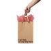 Hand holding a kraft gift bag with brown ribbon handles and pink tissue paper, printed with "I got this on my way here" text.