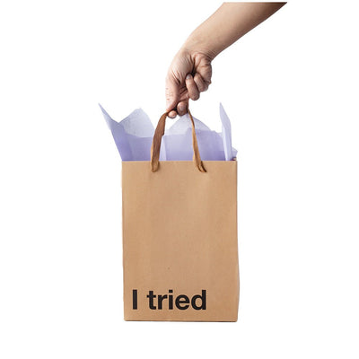 Hand holding a kraft gift bag with brown ribbon handles and "I tried" printed on the front, filled with white tissue paper.