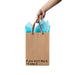 Hand holding a kraft gift bag with brown ribbon handles and blue tissue paper, printed with "If you don't like it, I'll take it" text.