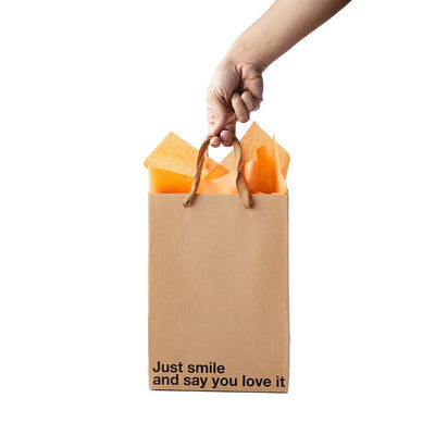 Hand holding a Kraft gift bag with brown ribbon handles and orange tissue paper, printed with "Just smile and say you love it" text.