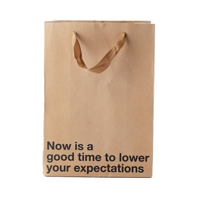 Brown kraft gift bag with brown ribbon handles and black text reading Now is a good time to lower your expectations
