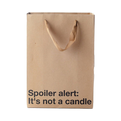 Kraft paper gift bag with brown ribbon handles and black text reading "Spoiler alert: It's not a candle" on front.