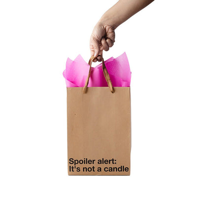 Hand holding a Kraft gift bag with brown ribbon handles and pink tissue, text reads Spoiler alert: It's not a candle.