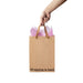 Hand holding a kraft gift bag with brown ribbon handles and pink tissue paper, printed with "Wrapping is hard" text.