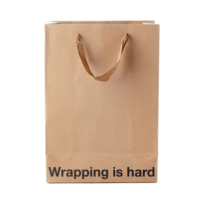 Kraft paper gift bag with brown ribbon handles and "Wrapping is hard" printed in black at the bottom front.