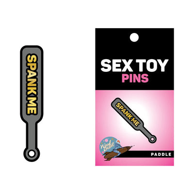 Gray enamel paddle pin with gold "SPANK ME" text, displayed on a pink and black Wood Rocket Sex Toy Pins backing card.