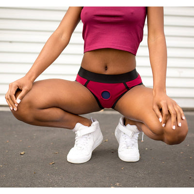 Woman wearing a red and black EmEx Contour Harness with white sneakers, squatting outdoors against a white wall background.