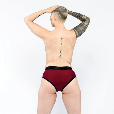 Person with short hair and tattooed arm wearing burgundy and black EmEx Contour Harness underwear, shown from the back.