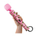 Hand holding a pink wand massager with green weed leaf pattern and a black power cord attached.