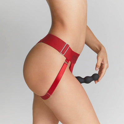 Side view of a person wearing a red adjustable strap-on harness with a black textured attachment against a neutral background.