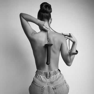 Black and white image of a woman with a high bun holding a leash attached to a dildo ring hanging down her back, wearing jeans.
