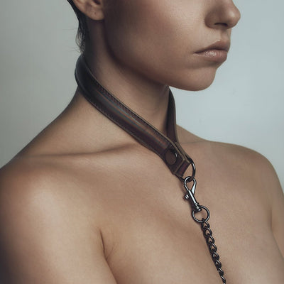 Close-up of a person wearing a dark leather leash collar with metal clasp and chain detail against a neutral background.