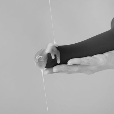 Black semi-realistic silicone dildo held in hand with liquid squirting from the tip, shown in monochrome.