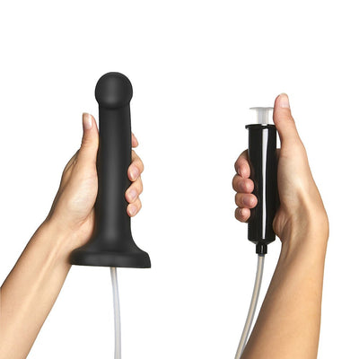 Hands holding a black semi-realistic silicone dildo connected to a syringe with a clear tube for squirting fluid.