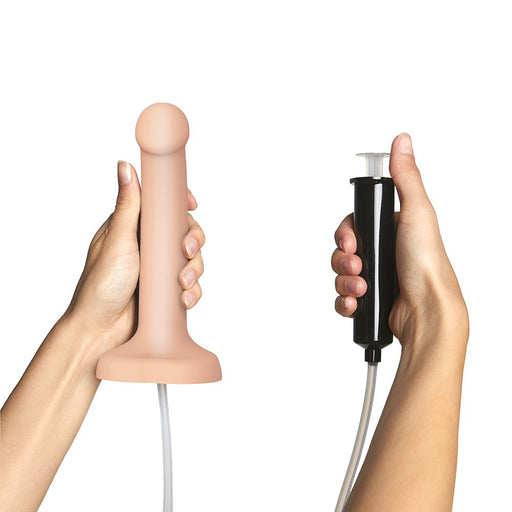 Hands holding a vanilla-colored semi-realistic silicone dildo connected to a black syringe for squirting fluid.