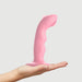 Hand holding a pink curved silicone dildo with a suction base and textured tapping tip against a plain background.