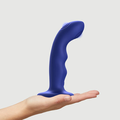 Hand holding a curved, blue silicone tapping dildo with a suction cup base on a light background.
