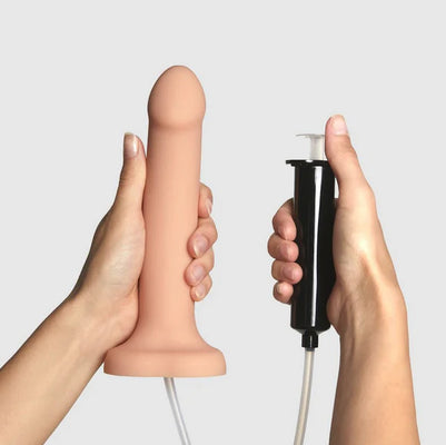 Hands holding a flesh-tone silicone dildo with attached tubing and a black syringe pump for fluid release.