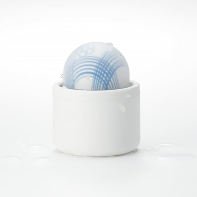Compact Tenga Iroha Temari Mizu in white holder with blue wave design, shown with water droplets on a white surface.
