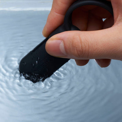 Hand holding a black silicone vibrating ring partially submerged in water, demonstrating its waterproof feature.