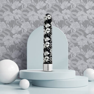 Black 7-inch vibrator with white floral pattern displayed on pastel blue circular platforms against a gray floral and snake wallpaper background.