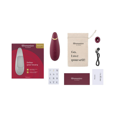 Womanizer Premium 2 clitoral stimulator in burgundy with packaging, USB charging cable, two stimulation heads, storage pouch, and user guides.