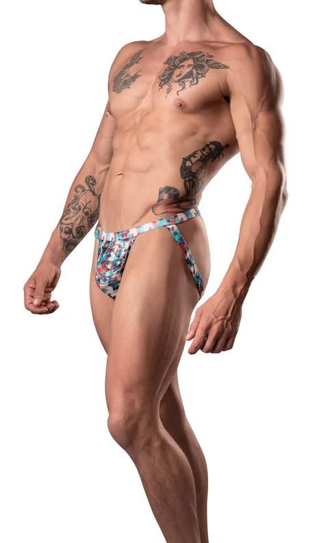 Male model wearing red, white, and blue low-cut jockstrap with contoured pouch and leg bands, showcasing athletic fit and tattoos.