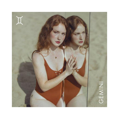 Red-haired woman in rust-colored swimsuit touching a mirror, Gemini zodiac symbol and text visible on sandy background.