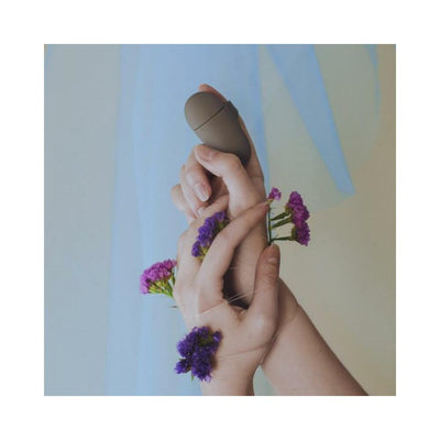 Hands wrapped with delicate purple flowers holding a sleek, taupe silicone finger vibrator against a soft blue background.